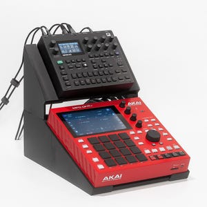 Puede incluir: Una configuración de estudio de producción de música electrónica en negro y rojo. La unidad roja, etiquetada como "MPC One+", presenta una pantalla táctil, botones y perillas. Una unidad negra se encuentra encima, conectada por cables. La configuración está sobre un soporte negro.
