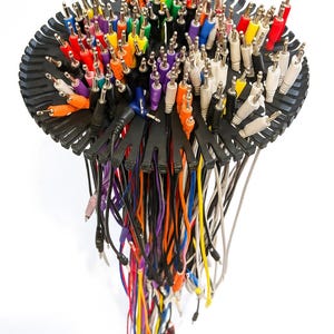 Pode incluir: Uma coleção de cabos e conectores de áudio coloridos, incluindo vermelho, amarelo, verde e azul, dispostos em uma forma abstrata. Os cabos estão presos a uma base preta circular, com os fios caindo para baixo.