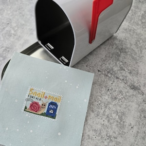 May include: A white mailbox with a red flag, open to reveal a light blue square with a cross-stitch design. The design features a snail, a mailbox, and the words "Snail Mail Forever 2124".