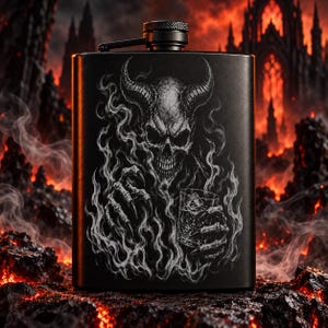May include: A matte black metal flask featuring a detailed white graphic of a horned skull figure with clawed hands holding a glass of liquid. The flask has a cylindrical screw-top cap with a textured grip and a hinged arm attachment at the top.