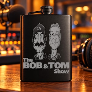 May include: A matte black metal flask featuring a laser-engraved caricature of two men, one wearing a baseball cap with the letters LA. Below the figures, the text reads The BOB & TOM Show in a bold, sans-serif font. The flask has a screw-top lid.