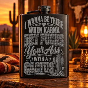 May include: A matte black metal flask featuring an engraved design with a cactus illustration and the text: I WANNA BE THERE WHEN KARMA DRY FUCKS YOUR ASS WITH A CACTUS. The flask has a screw-top cap with a textured grip and a hinged metal arm.