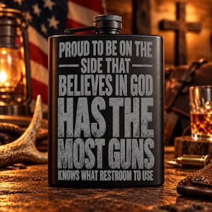May include: A matte black metal flask featuring the engraved text: PROUD TO BE ON THE SIDE THAT BELIEVES IN GOD HAS THE MOST GUNS KNOWS WHAT RESTROOM TO USE. The flask has a screw-top cap with a textured grip and a hinged arm attachment.