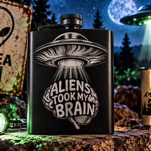 May include: A matte black metal flask featuring a white laser-engraved graphic of a UFO hovering over a human brain. The text "ALIENS TOOK MY BRAIN" is centred within the design. The flask has a screw-top cap and a rectangular, curved body shape.