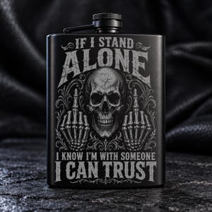 May include: A matte black metal flask featuring an engraved design of a human skull flanked by two skeletal hands making middle finger gestures. The text reads: IF I STAND ALONE I KNOW I'M WITH SOMEONE I CAN TRUST. It has a screw-top lid.