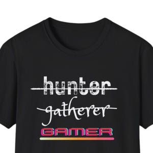 May include: Black t-shirt featuring the text "hunter" crossed out, then "gatherer", and "GAMER" in a colourful, gradient font. A casual wear item.