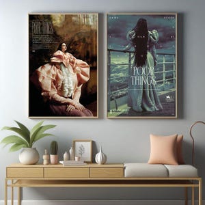 May include: Two framed movie posters. The left poster features a woman in a pink dress with the text "Poor Things". The right poster shows a woman in a long dress with the text "Poor Things". Both posters are in gold frames.