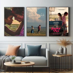 May include: Three framed movie posters for "Bones and All" are displayed above a gray sofa. The posters feature images of the film's stars and the movie title. The room is decorated with a round wooden coffee table and side tables.