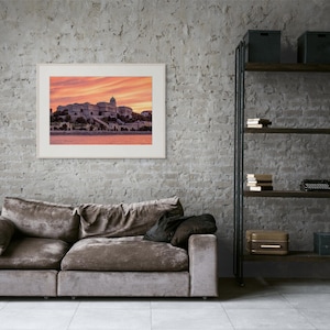 Può includere: Una stampa incorniciata di un paesaggio urbano al tramonto è appesa sopra un divano di velluto marrone. Una libreria in metallo nero con libri e oggetti decorativi si trova a destra. Il muro è di mattoni grigi.