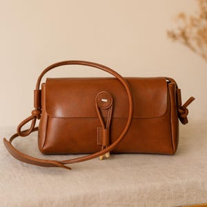 May include: A brown leather crossbody bag with a flap closure and a long, adjustable strap. The bag features a button closure and decorative knots on the strap attachments. The bag is rectangular and has a minimalist design.
