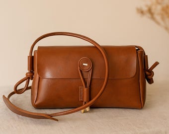 May include: A brown leather crossbody bag with a flap closure and a long, adjustable strap. The bag features a button closure and decorative knots on the strap attachments. The bag is rectangular and has a minimalist design.