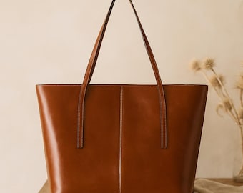 May include: A brown leather tote bag with long handles. The bag has a rectangular shape with a vertical seam down the center. The bag is sitting on a wooden surface.