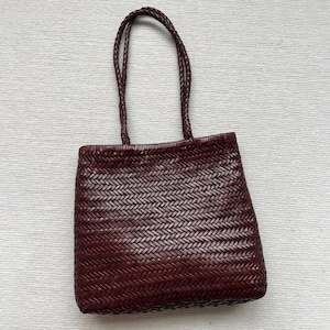 May include: A burgundy-colored tote bag with a woven design. The bag has two braided handles and a rectangular shape. The weave pattern is visible across the entire surface of the bag, creating a textured appearance. The handles are also braided.