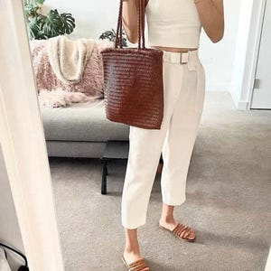 May include: A dark brown woven leather tote bag with two braided shoulder straps, paired with a white spaghetti strap crop top, white high-waisted trousers with a matching belt, and brown braided leather slide sandals.