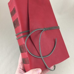 May include: A red leather journal with a wrap-around closure. The spine has decorative stitching with dark thread. A grey leather cord wraps around the journal to secure it. The journal is held in a hand.