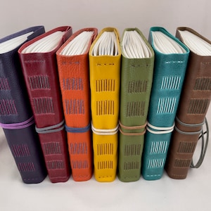 May include: Seven leather-bound journals in various colors, including purple, red, orange, yellow, green, teal, and brown. Each journal has a band around it and is stitched with a contrasting thread.