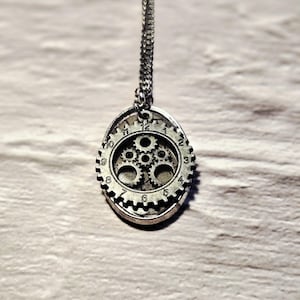 May include: Silver steampunk pendant necklace. The oval pendant has a clock face design with visible gears and numbers 1 to 12. The necklace chain is silver and is attached to the top of the pendant.