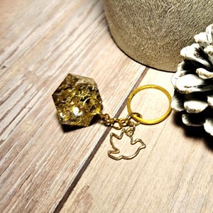 May include: A gold-colored keychain with a hexagonal resin charm filled with gold flakes. The keychain features a gold ring and a gold dove-shaped charm. The background includes a wooden surface, a textured pot, and a pinecone.