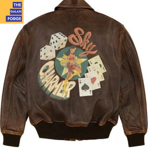 May include: Brown leather bomber jacket with a vintage-style graphic on the back. The graphic features dice, playing cards, and a pin-up girl with the words "Sky Charmer". The jacket has a ribbed collar and cuffs.