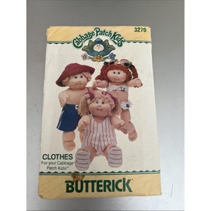 Op de afbeelding: Een vintage Butterick-patroon voor Cabbage Patch Kids-kleding. Het patroon toont illustraties van drie Cabbage Patch Kids-poppen in badkleding en casual outfits. De verpakking is crèmekleurig met het Cabbage Patch Kids-logo en het woord "CLOTHES".