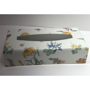 May include: A rectangular tissue box featuring a floral design. The box is white with a grey oval opening and is adorned with colourful flowers in shades of yellow, blue, and pink. The design includes various floral and leaf motifs.