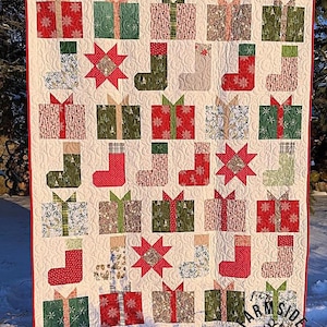 Può includere: Una trapunta natalizia festosa con un motivo ripetuto di scatole regalo, calze e forme di stelle in tessuti rossi, verdi e fantasia. La trapunta ha uno sfondo color crema e un bordo rosso. Il testo "Farmside Quilting" è visibile in basso a destra.