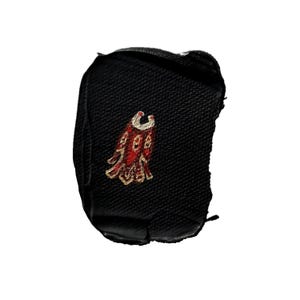 May include: A black fabric patch with a red and gold embroidered design. The design appears to be a stylized bird or emblem. The patch is rectangular with rounded edges.