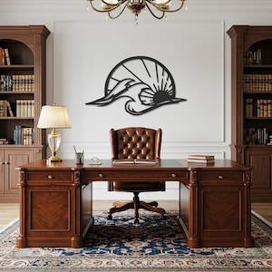 May include: A black metal wall art piece depicting a stylized seagull soaring over waves with a sun in the background. The artwork is mounted on a white wall above a wooden desk and leather chair in a home office setting.