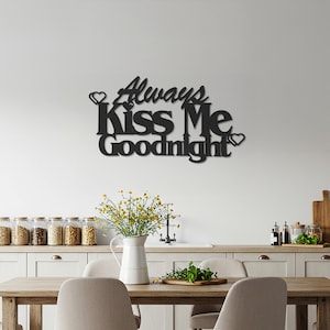May include: Black metal wall art with the words "Always Kiss Me Goodnight" in a decorative font. The sign includes heart accents. The art is mounted on a white wall above a dining table with a vase of flowers.