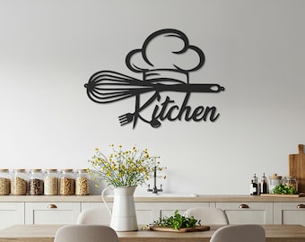 May include: Black metal kitchen wall art featuring a chef's hat, whisk, and the word "Kitchen" in a decorative font. The art piece is mounted on a white wall above a kitchen table and cabinets.