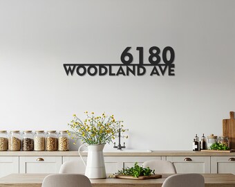 May include: Black metal house number sign with the address "6180 WOODLAND AVE" mounted on a white wall. The sign is above a wooden table with a white pitcher of yellow flowers.