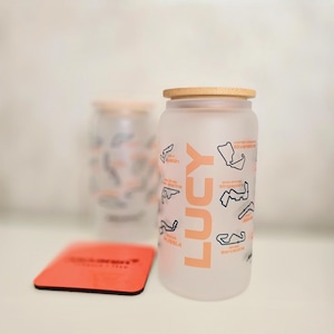 May include: A frosted glass tumbler with a bamboo lid, featuring the name LUCY in large orange letters and various black outlines of Formula 1 race tracks with orange text labels. A square orange coaster sits in the foreground next to the glass.