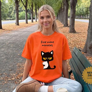 May include: A bright orange short-sleeved t-shirt featuring a graphic of a black cat holding a white pillow with the text Five more minutes printed in black above the cat. The shirt has a crew neckline and a relaxed fit.