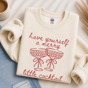May include: A cream-colored crewneck sweatshirt featuring a red graphic print of two stemmed cocktail glasses with bows. The text reads, "have yourself a merry little cocktail" in a cursive font. The garment has long sleeves and a ribbed collar.