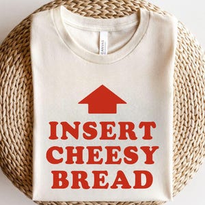 May include: Cream-colored t-shirt with the text "INSERT CHEESY BREAD" in bold red letters, topped with a red arrow pointing upwards. The shirt is made of a soft material and is displayed on a woven surface.