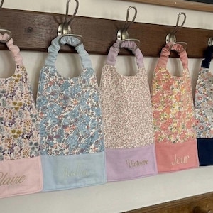 Peut inclure: Six bavoirs à motifs floraux suspendus à un porte-manteau en bois. Chaque bavoir présente un motif floral différent et un bas uni avec des noms brodés. Les bavoirs sont dans les tons de rose, de bleu et d'orange.
