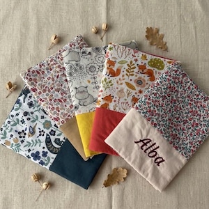 Peut inclure: Cinq pochettes en tissu rectangulaires disposées en éventail. Chaque pochette présente une section supérieure à motifs et une section inférieure unie. Une pochette crème porte le prénom Alba brodé en écriture cursive marron foncé.