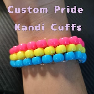 May include: A beaded Kandi cuff bracelet in the colors of the Pansexual Pride flag: pink, yellow, and blue. The bracelet is made of small, round beads strung together. Text on the image reads "Custom Pride Kandi Cuffs."