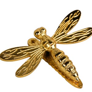 Puede incluir: Figura de libélula dorada con alas y cuerpo detallados. La libélula es de metal y tiene un acabado brillante. Las alas están extendidas y el cuerpo segmentado. La figura es un artículo decorativo.