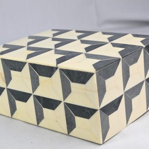 Può includere: Una scatola rettangolare con un motivo geometrico. La scatola presenta una base color crema con un motivo ripetuto di forme grigio scuro. Il design crea un effetto visivo sorprendente, adatto per riporre piccoli oggetti.