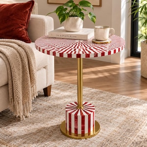 May include: A round side table featuring a red and white sunburst pattern on the tabletop and base, connected by a central gold-toned metal pole. The table surface holds a white book titled The Kinfolk Home, a small potted plant, and a mug.