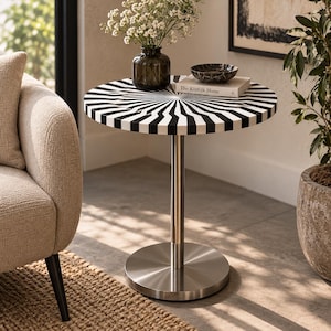 May include: A round side table featuring a black and white sunburst pattern on the tabletop, supported by a central silver-toned metal pedestal base. The table holds a dark glass vase with white flowers, a black marble bowl, and a book titled The Kinfolk Home.