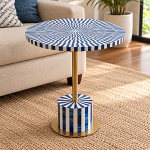 May include: A round side table featuring a blue and white sunburst pattern on the tabletop and a matching cylindrical base. The table is supported by a central gold-toned metal pole and rests on a circular gold-toned metal base plate.