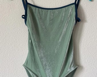 May include: A sage green velvet bodysuit featuring a square neckline and dark navy blue spaghetti straps. The garment has a high-cut leg opening and a soft, shimmering texture throughout the fabric, suspended on a light blue plastic hanger.