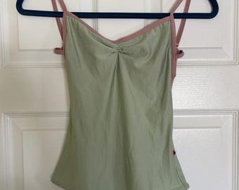 May include: A sage green leotard featuring a sweetheart neckline with a central twist detail and thin, dusty pink straps. The garment has a smooth, solid-coloured fabric construction with a high-cut leg opening and a small red tag on the side.