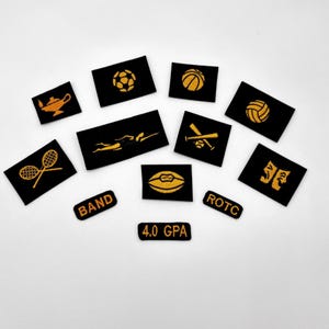 Może przedstawiać: Kolekcja czarnych, haftowanych naszywek ze złotymi wzorami. Naszywki przedstawiają różne sprzęty sportowe, pływaka, lampę dżina oraz napisy "BAND", "ROTC" i "4.0 GPA".