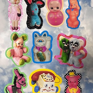 May include: A collection of nine stickers featuring various vintage-style toys, including a doll in a pink dress, a blue rabbit, a pink round doll, an orange poodle, a pink bear-costumed doll, a white cat figurine, two furry creatures, a pink donkey, a rotary phone, and a pink bear.