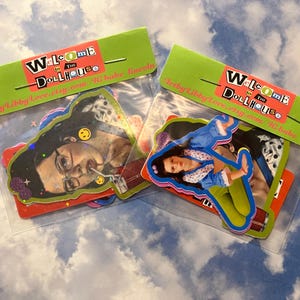 May include: Two clear plastic bags containing colorful stickers. Each bag has a green header with the words "Welcome to the Dollhouse" in a mix of fonts and colors. The stickers feature images of people and smiley faces.