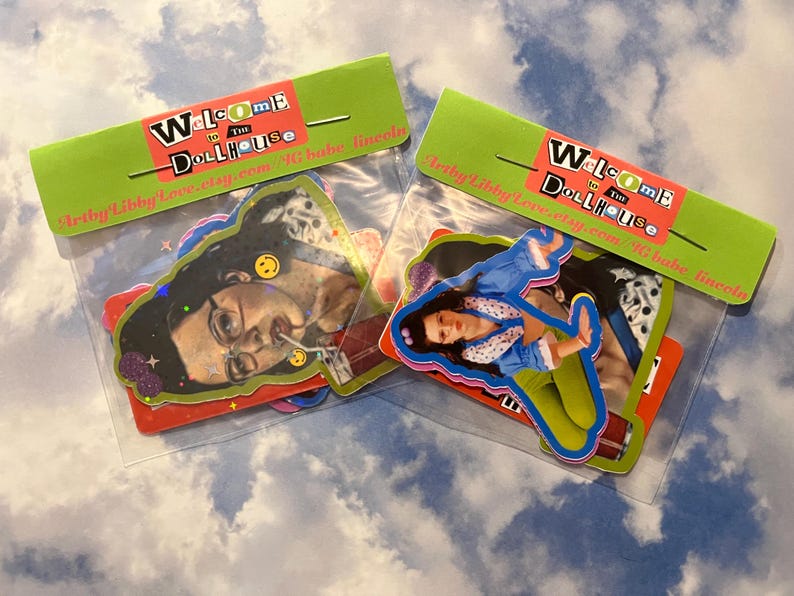 May include: Two clear plastic bags containing colorful stickers. Each bag has a green header with the words "Welcome to the Dollhouse" in a mix of fonts and colors. The stickers feature images of people and smiley faces.