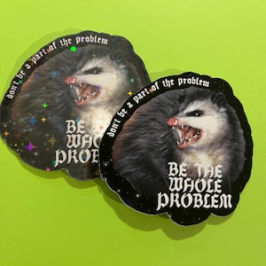 Könnte beinhalten: Zwei Aufkleber mit schwarzem Rand und einer Opossum-Illustration. Das Opossum hat den Mund geöffnet und zeigt seine Zähne. Der Text auf den Aufklebern lautet "don't be a part of the problem" und "BE THE WHOLE PROBLEM."